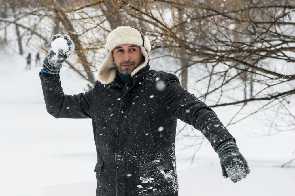 A man in winter clothing enjoys a snowy day, playfully throwing a snowball outdoors.