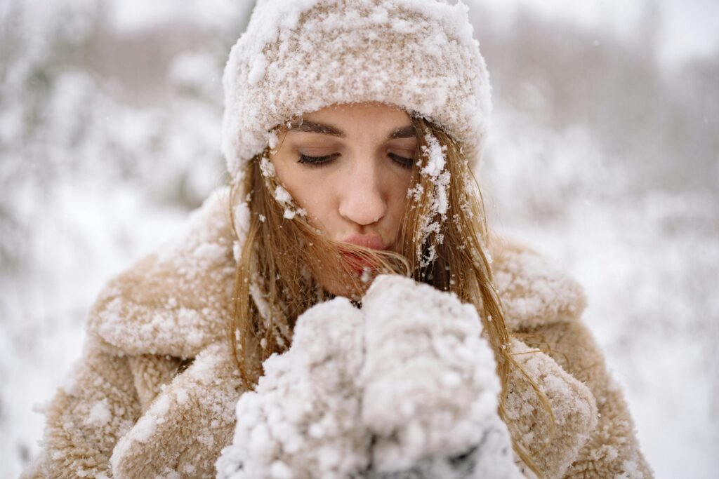 Close-up of a woman in winter attire surrounded by snow, blowing warm air into her hands.