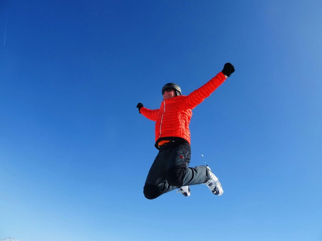 A joyful man leaps midair against a clear blue sky, wearing ski gear in a winter setting.